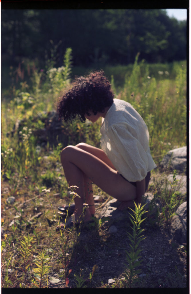 Frau ist in der Hocke und steht gerade auf, trägt Sommerkleidung auf einer Wiese