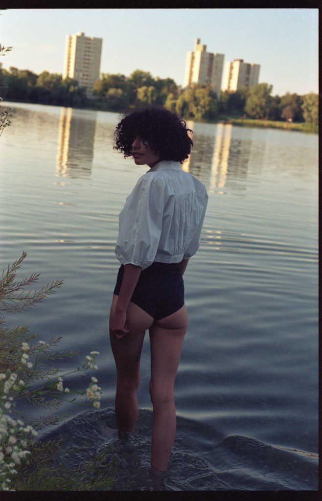 analoge Fotografie von einem weiblichen Model, dass in einen See steigt und zurück über ihre Schulter guckt. Im Hintergrund ist ein hochhaus zu sehen. Sehr urban.
