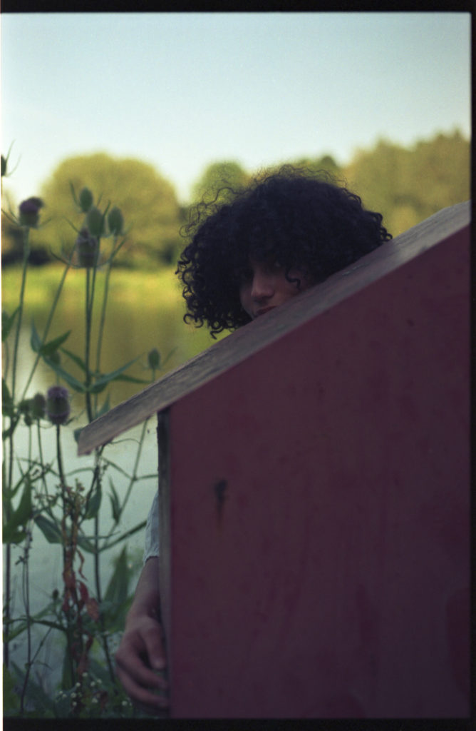 Eine Junge Frau schaut hinter einem Kasten an einem See hervor. Das Foto ist sehr künstlerisch.