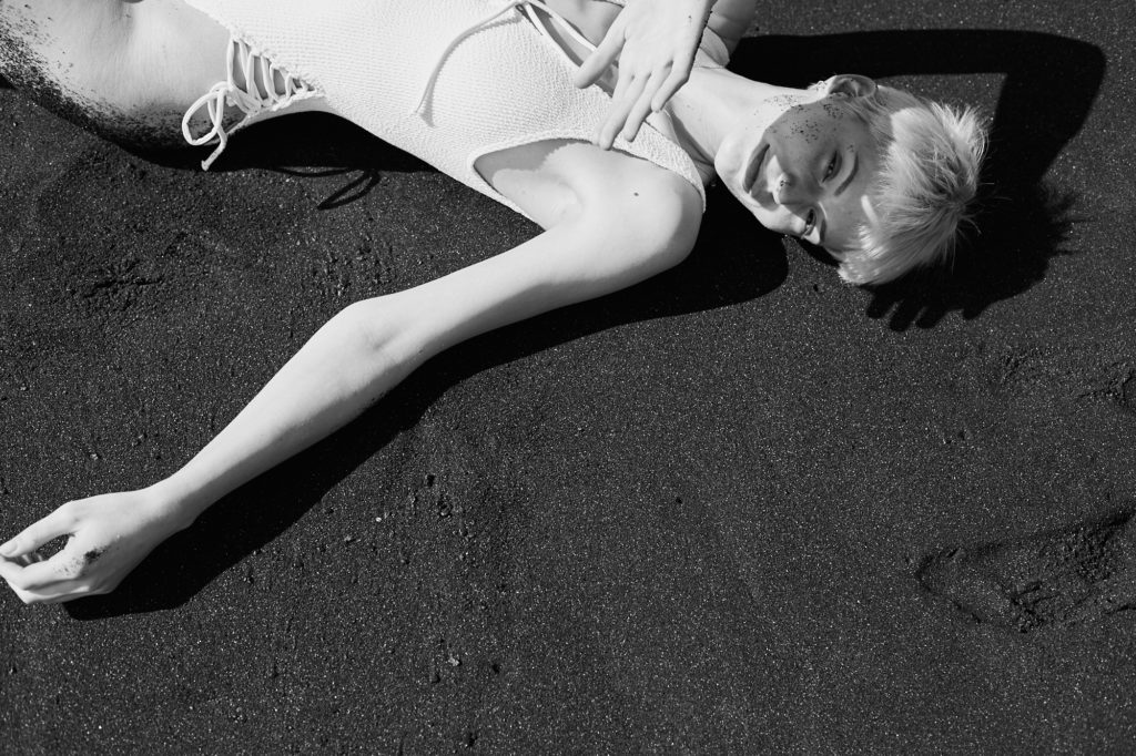 Eine schwarz weiß Fotografie von einem weiblichen Model mit kurzen blonden Haaren im schwarzen Sand von oben fotografiert. Sehr stark durch die schwarz weiß Kontraste.