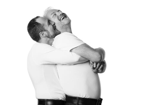 männliches paar, einer umarmt den anderen von hinten, sie lachen und sehen sehr natürlich verliebt aus.