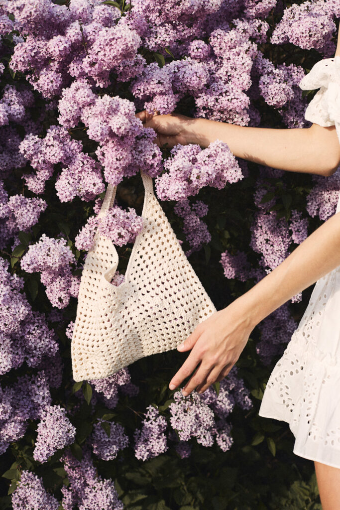 Ein Foto von einer jungen Frau, die eine Handtasche vor einem Fliederbusch hält.