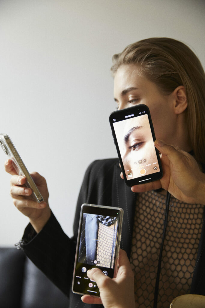 ein weibliches Model hält ein Handy in der Hand und wird während dessen von anderen Handys fotografiert. Moderne Modefotografie.