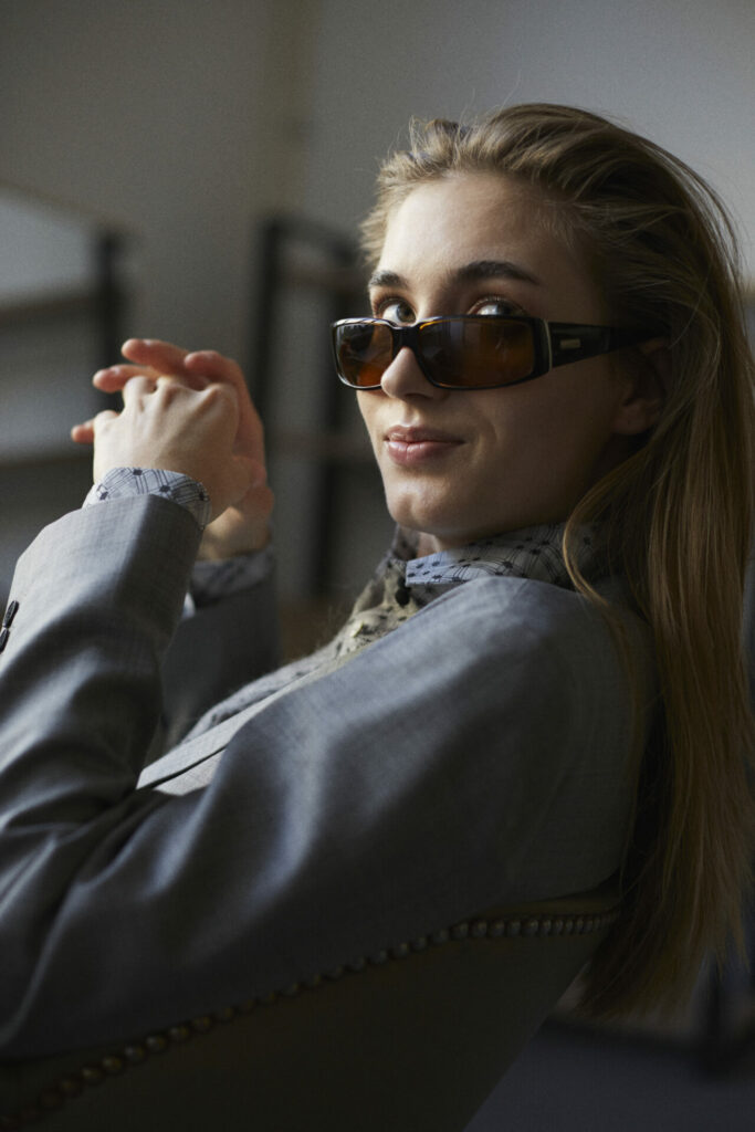 Weibliches Model mit grauem Blazer und Sonnenbrille, schaut frech über die Kamera hinweg.