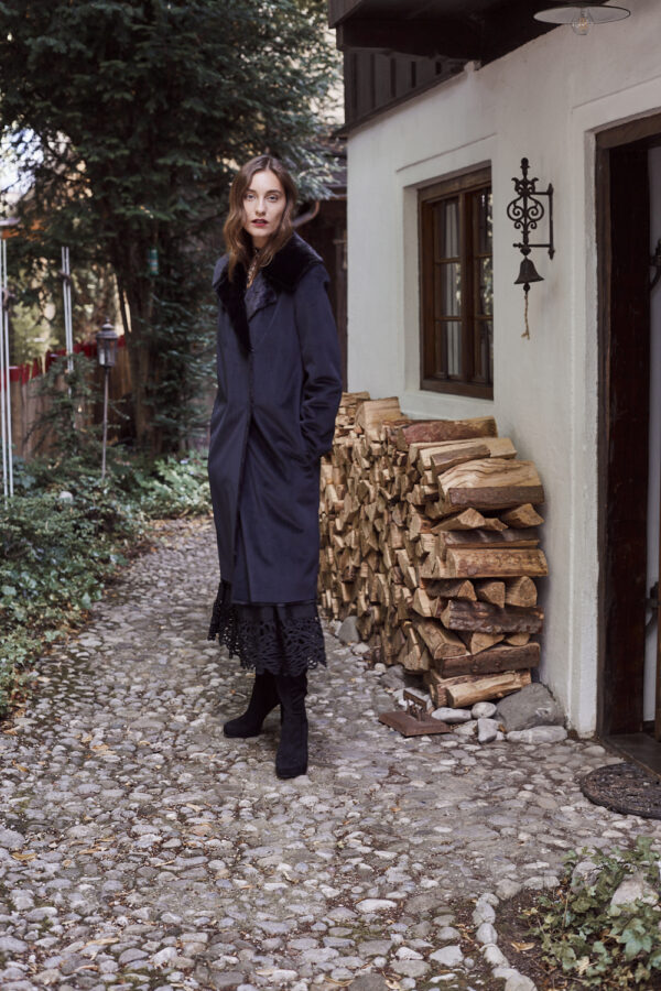 Ein Model mit Wintermantel steht vor einem alten Holzhaus.