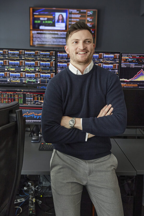 Ein junger Mann steht lachend vor einem Display mit vielen Aktien und Zahlen.