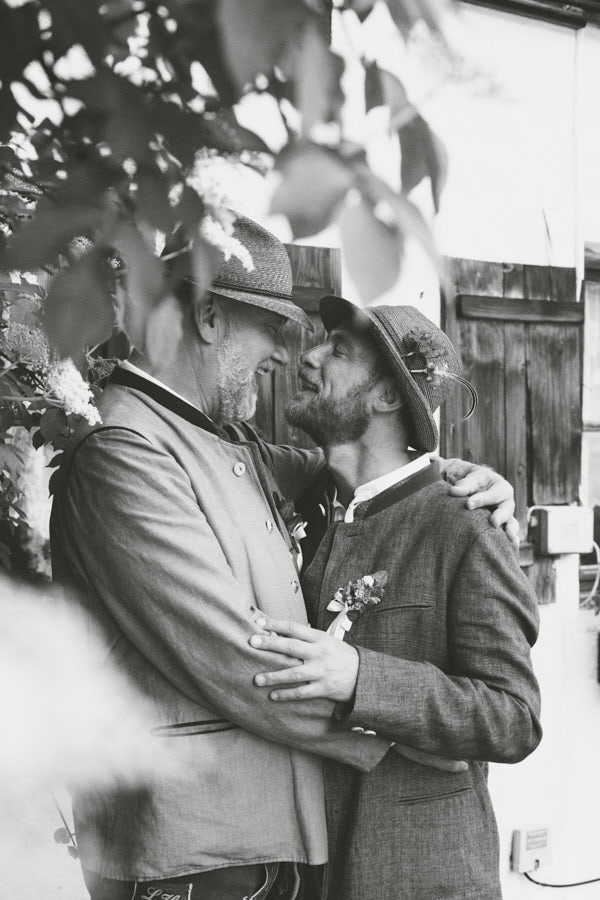Ein schwarz weiß Foto von einem schwulen Hochzeitspaar in Tracht, dass sich umarmt und anlacht.