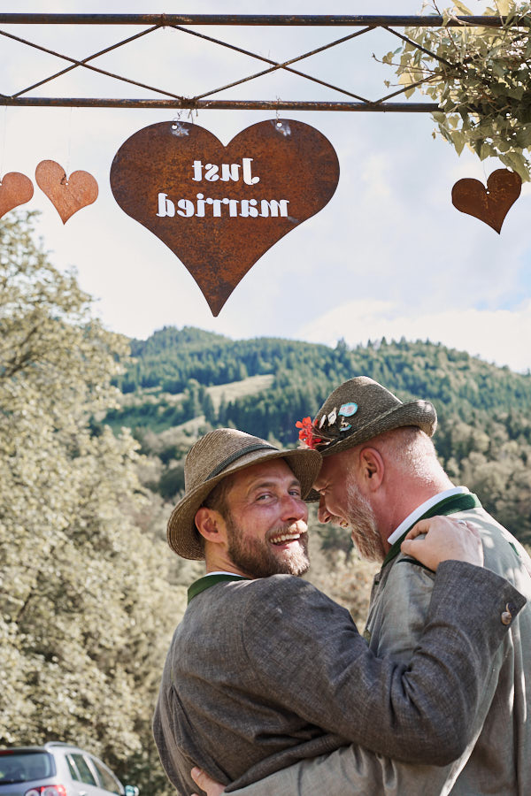 Schwules Hochzeitspaar in Tracht, die sich umarmen und zurück schauen in die Kamera. Über Ihnen steht auf einem Metallschild