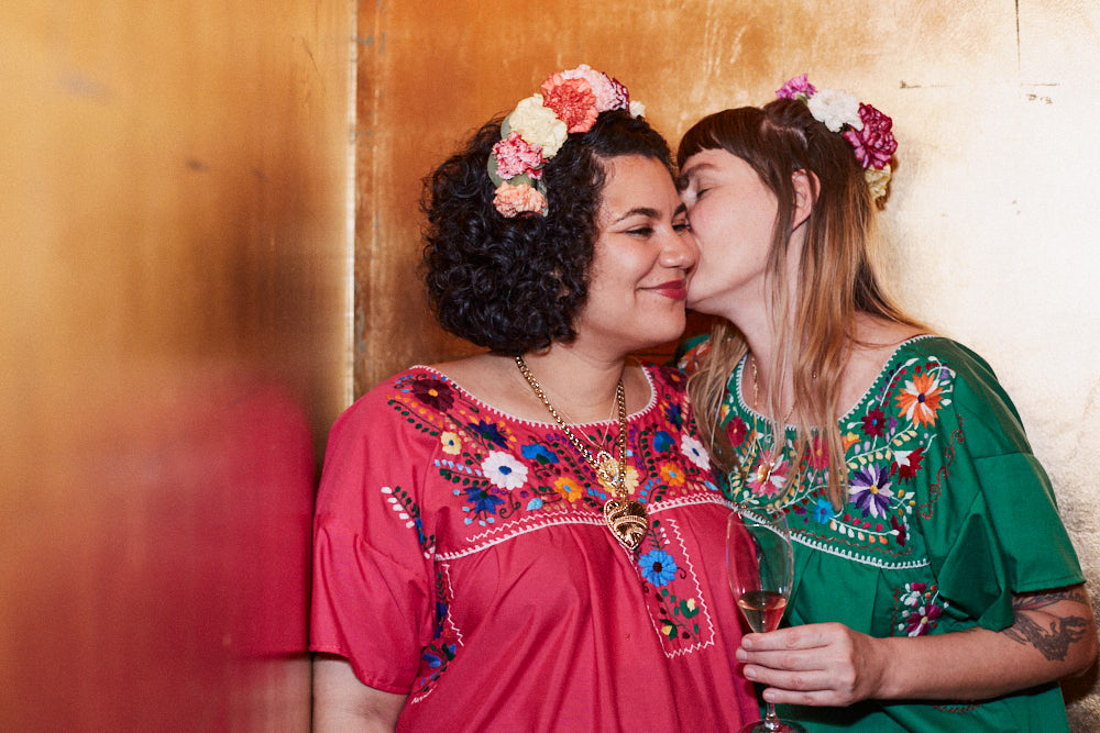 Zwei Frauen vor einer goldenen Wand in bunten Kleidern, die sich Küssen.